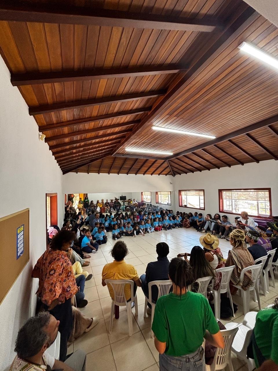 Salão amplo cheio de crianças, educadores e famílias reunidos durante uma atividade da Pequena Tribo.