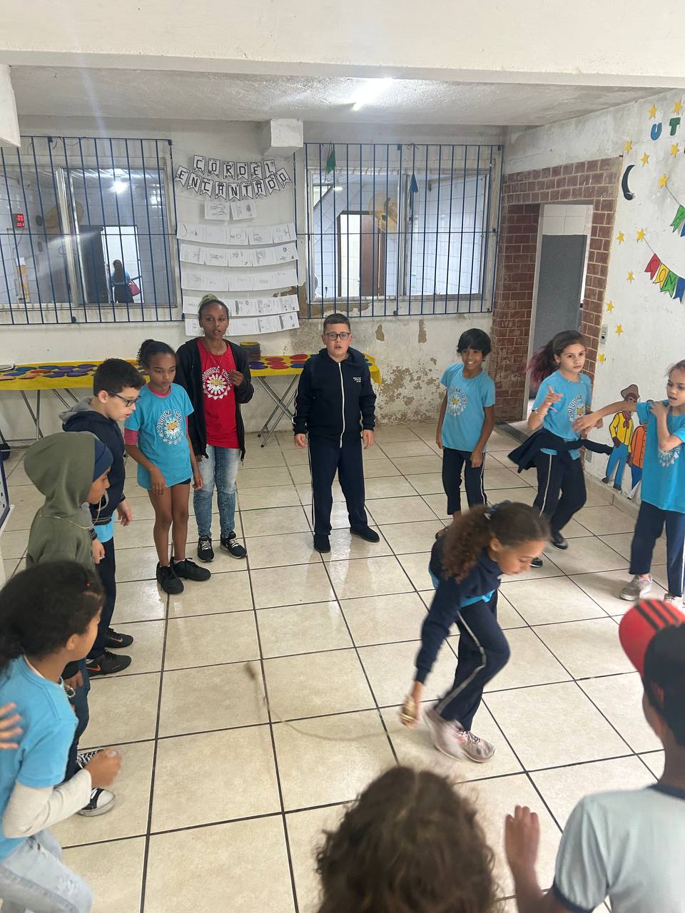 Crianças em roda numa sala, uma delas se movimentando no centro, durante brincadeira de recreação.