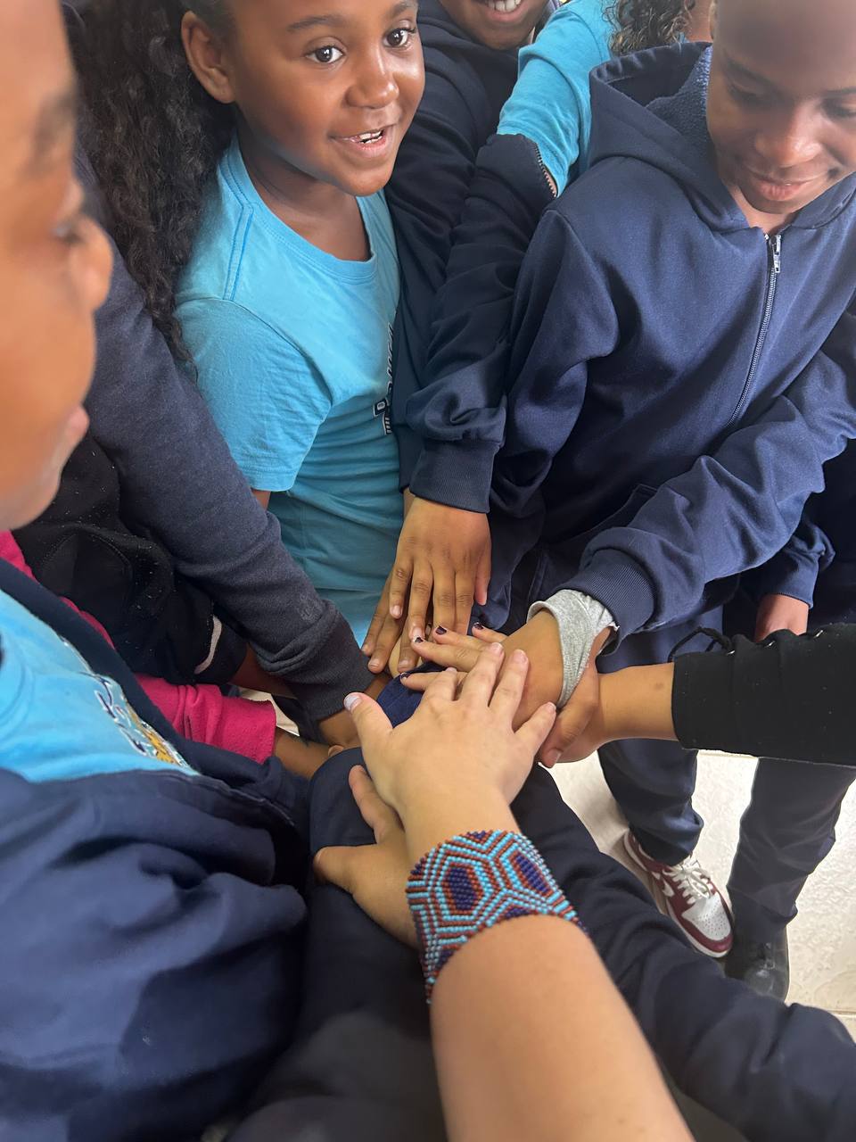 Crianças e uma educadora com as mãos sobrepostas em círculo, em gesto de união.